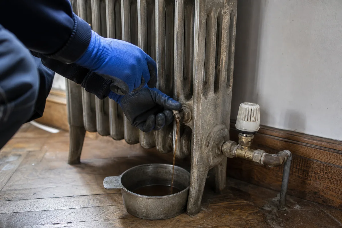Mains gantées purgeant un radiateur en fonte dans un salon de maison en brique rouge